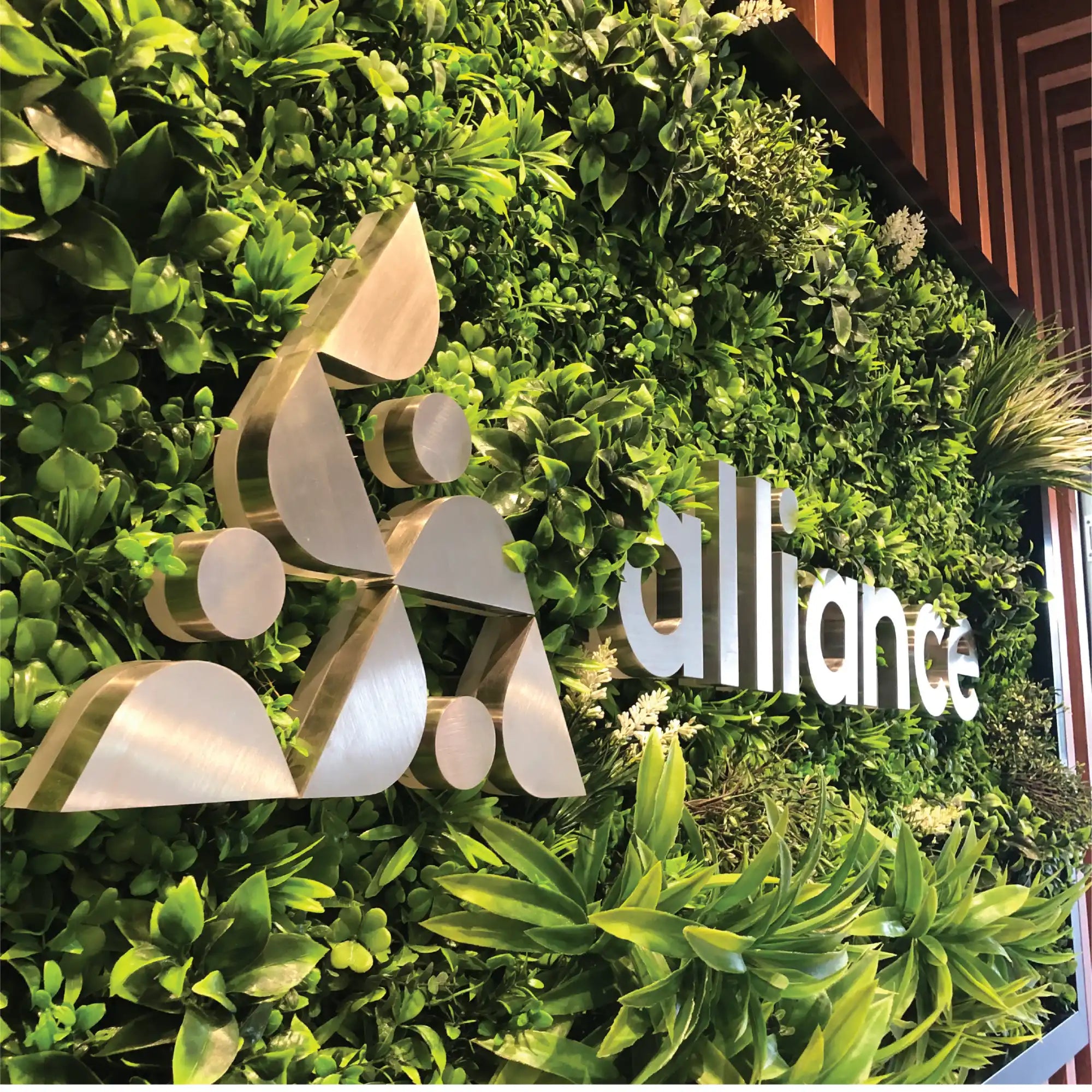 A metallic ’Alliance’ sign mounted on a vibrant green wall of plants.
