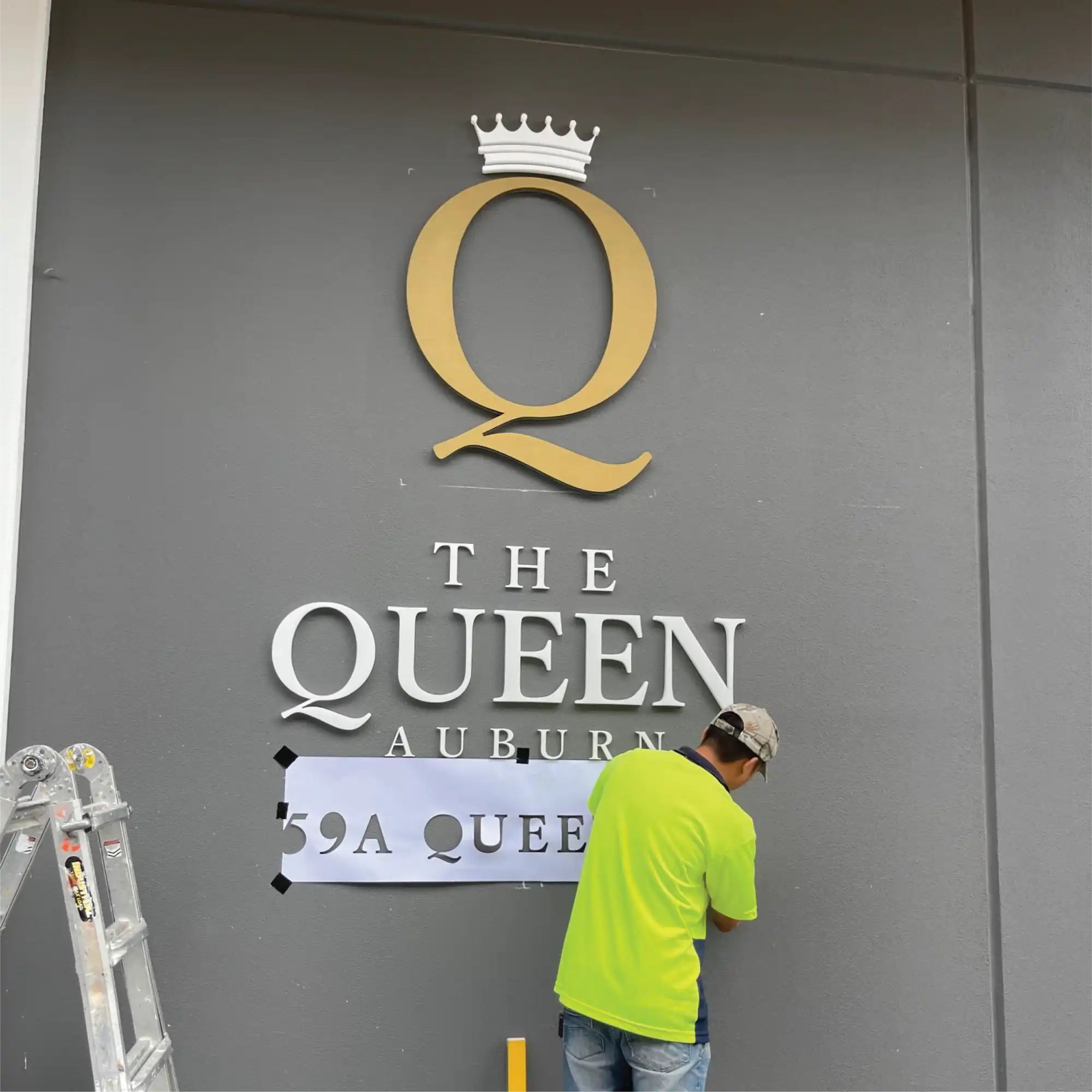 A building sign with a large, golden letter ’Q’ wearing a crown, above the words ’THE QUEEN AUBURN’ in silver lettering.